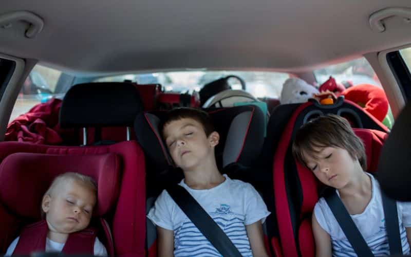 Deux enfants et un bébé à l'arrière d'une voiture, tous endormis. On voit des affaires dans le coffre derrière eux.