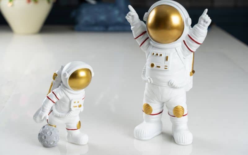 Deux figurines de cosmonautes blanches et dorées. Une petite et une plus grande.